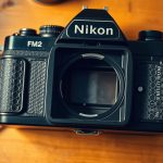 Nikon FM2 SLR camera body with back plate open showing mirror mechanism and prism chamber on wooden workbench with precision repair tools